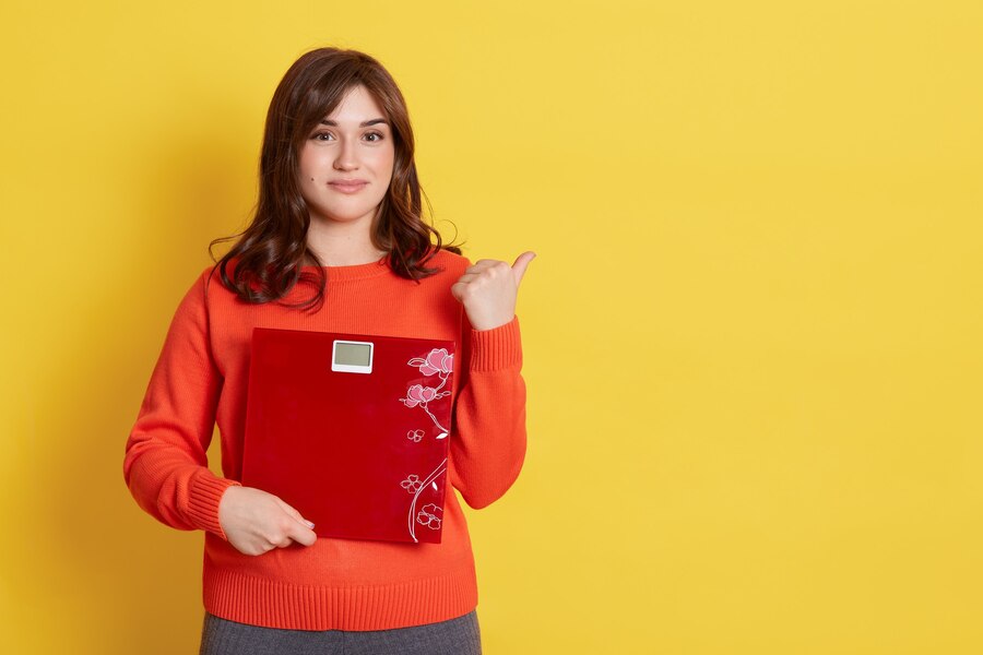 female with pleasant appearance dark hair holding floor scales hands has calm expression pointing away with finger copy space isolated yellow wall