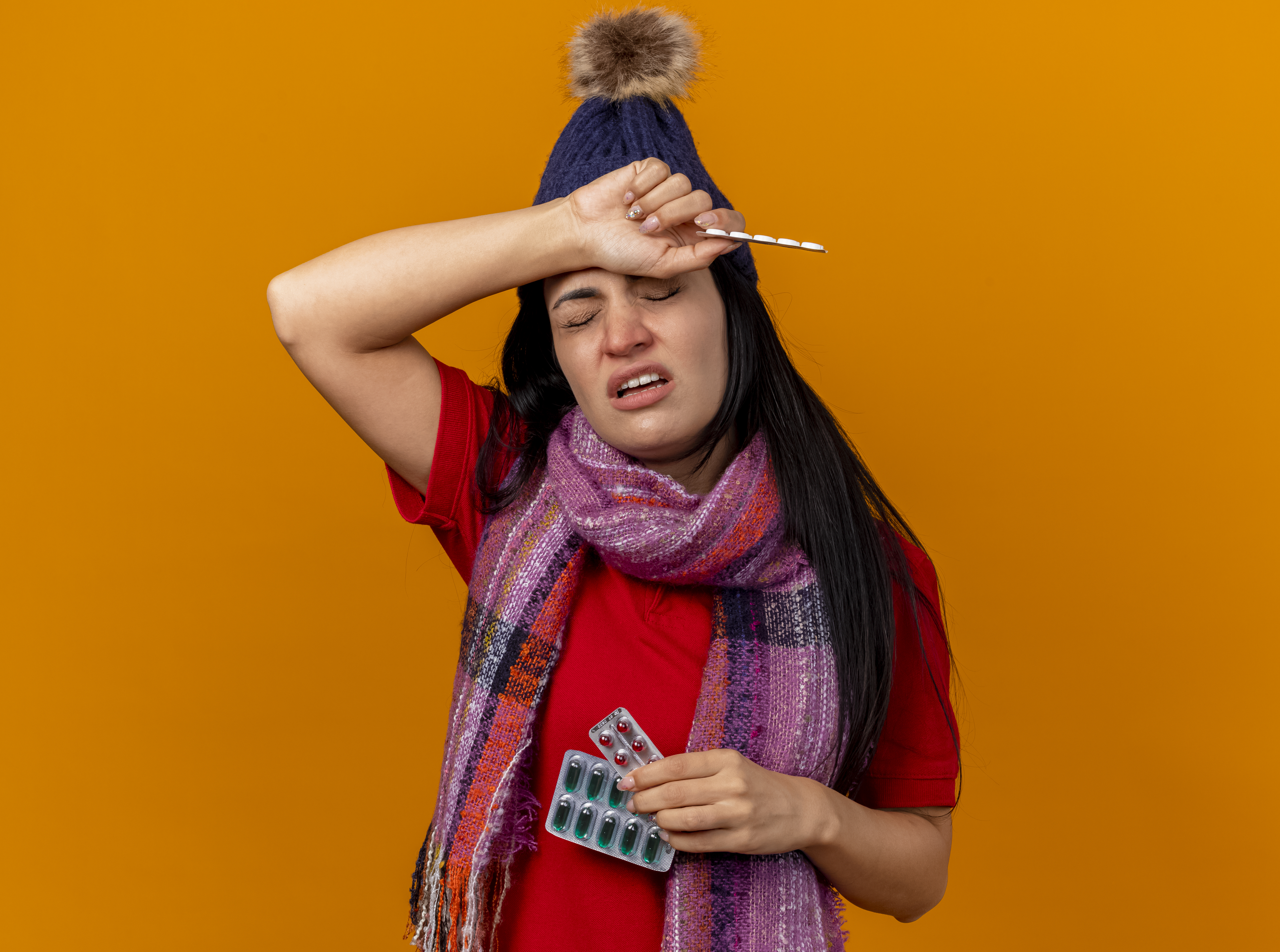aching-young-caucasian-ill-girl-wearing-winter-hat-scarf-holding-packs-capsules-tablets-putting-hand-head-with-closed-eyes-isolated-orange-wall-with-copy-space
