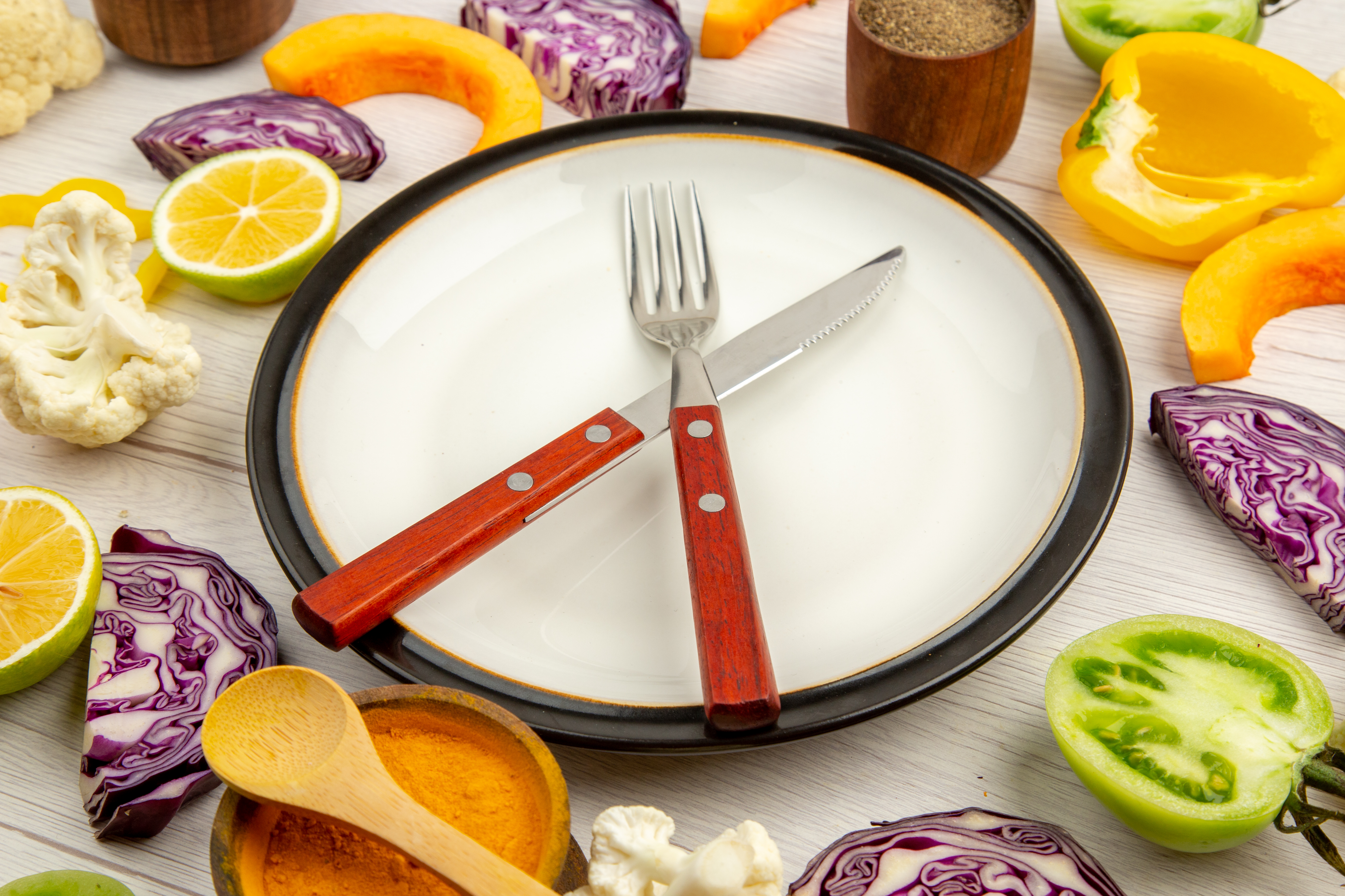 bottom-view-cut-vegetables-pumpkin-red-cabbage-lemon-green-tomatoes-cauliflower-yellow-bell-peppers-crossed-knife-fork-plate-spices-small-bowls-table
